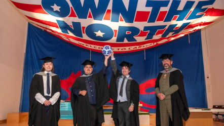 Four males in academic robes with two in the middle holding aloft a football and stood underneath a huge flag emblazoned with Own The North