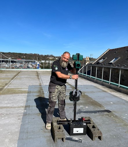 A man stands on a roof holding a pole with a box on top. The sky is blue the man is smiling at the camera.