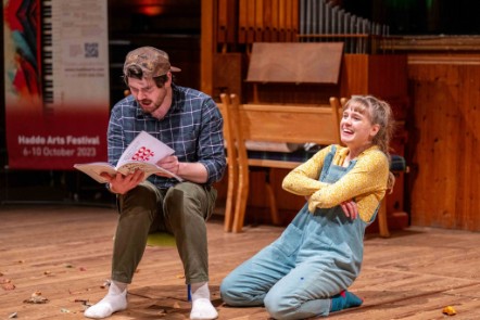 Two stage performers. A boy with a cap on back to front is reading a magazine and sitting on a small chair, the girl is on the floor smiling