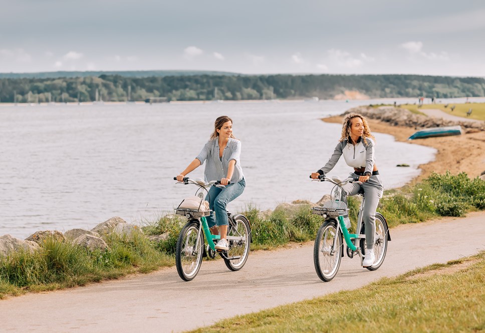Two female riders using Beryl bikes along a coastal path. Both riders are looking to their left at something off camera.