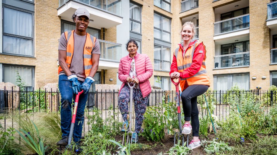 Three people stand with garden forks in a newly planted garden in front of a block of flats.