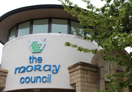 The exterior wall of Moray Council's headquarters, the writing is blue and there is a green bush in the background.