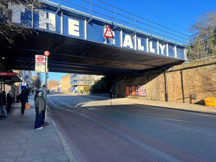 Two people wait at a bus stop on Caledonian Road, underneath 'The Cally' bridge