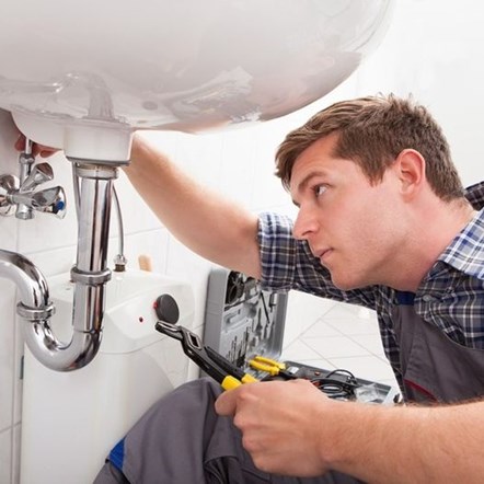 A man wearing a plumber's grey work overalls is under a white ceramic sink with a yellow and black tool and a grey toolbox beside him.