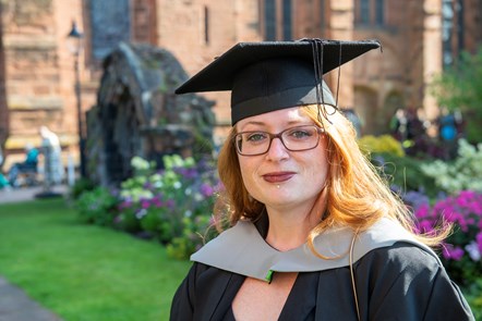 Female wearing glasses and black graduation academic gown and cap standing in front of grass, flowers and sandstone building