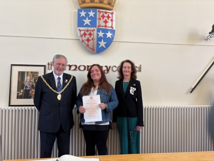 Three people stand in a line under a coat of arms. The one in the middle holds a certificate. They are all smiling.