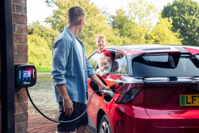 Motability Operations EV and customer: Man plugs in red electric vehicle using Ohme charge point and smiles at children sitting in the car