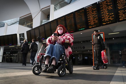 A young woman with pink hair sitting in a wheelchair in front of the departure screens at Birmingham New Street station, uses her phone to send a message.