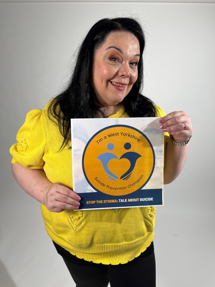 Suicide Prevention Champions - Lisa Riley B: Lisa is pictured smiling at the camera, holding the 'I'm a West Yorkshire Suicide Prevention Champion' logo on a piece of paper.