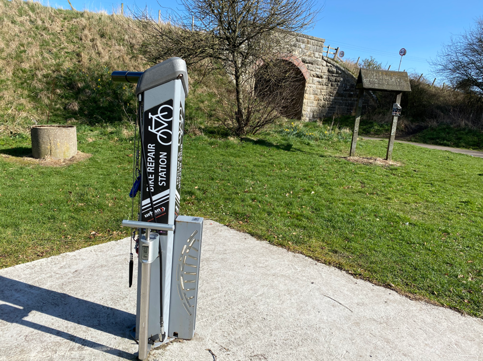 A metal bike repair station stands on a concrete base with a grass bank and a bridge arch in backgroud,  The sky is blue and it is summer.