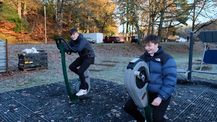 Two senior High School pupils are on exercise equipment at an outdoor gym, They are wearing warm jackets and smiling at the camera.