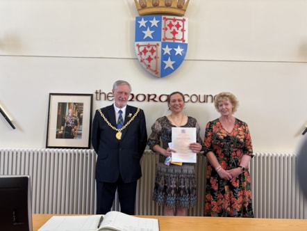 Three people stand in a line smiling at the camera. The one in the middle holds a certificate. There is a coat of arms above them.