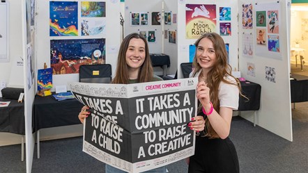 Two females at University of Cumbria Creative Community art exhibition with a newspaper. Headline 'It Takes a Community To Raise A Creative'