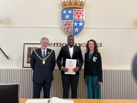 Three people stand in a line smiling at the camera. The person in the middle holds a certificate. There is a coat of arms above them.