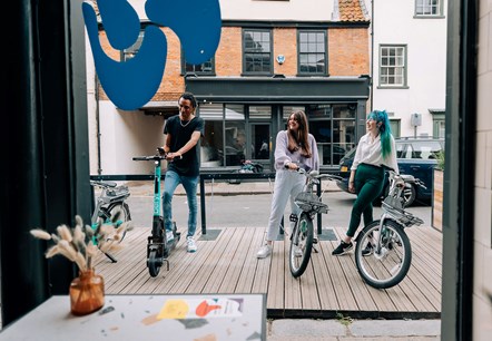 Three young people outside of a shop. One is boarding a scooter and two are stood by a Beryl bike.