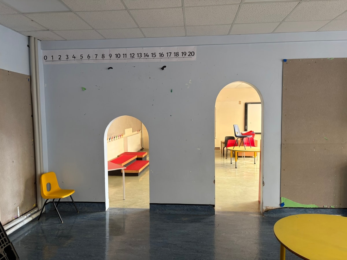 A view of a classroom with two arched doorways leading to another room, with various school furniture items