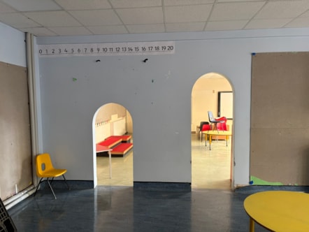 A view of a classroom with two arched doorways leading to another room, with various school furniture items