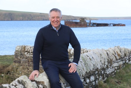 A man sits on a stone wall in front of the sea. A shipwreck is in the background.