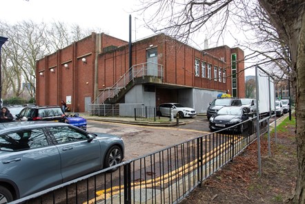 A low rise red brick building with few windows, metal stairs running up one side and metal fire doors, with cars parked outside.