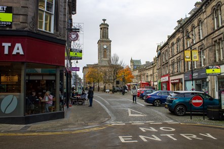 Shops on a high street have to let signs above them. People are walking on cobbles. Cars are parked on the right facing the shops.