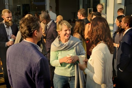 A group of people wearing a mix of smart business attire and casual clothing stand chatting in an outdoor area holding cups of coffee from a vending machine. A woman wearing a grey shawl, jeans, and green pullover is smiling as she chats to another woman at the forefront of the image.