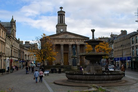 Two women walk down cobbled street past the fountain on Elgin High Street. The weather is overcast and it is a dull day.