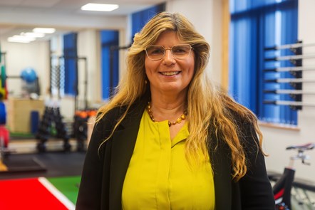 Kathryn Wain smiling to camera and wearing a bright green dress and black jacket stood against a backdrop of weight and gym equipment