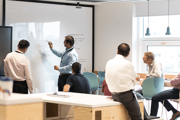 half year report new image: Five businessmen in office gather around a whiteboard, where another man stands writing on it