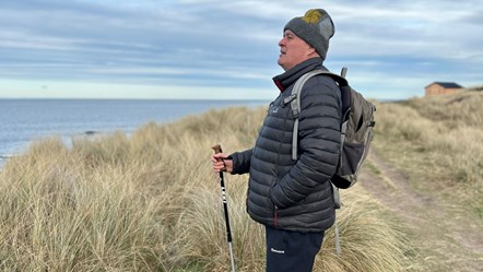 A man with a warm hat and black insulated jacket and backpack and hols a hiking pole as he looks out from sand dunes to the sea.