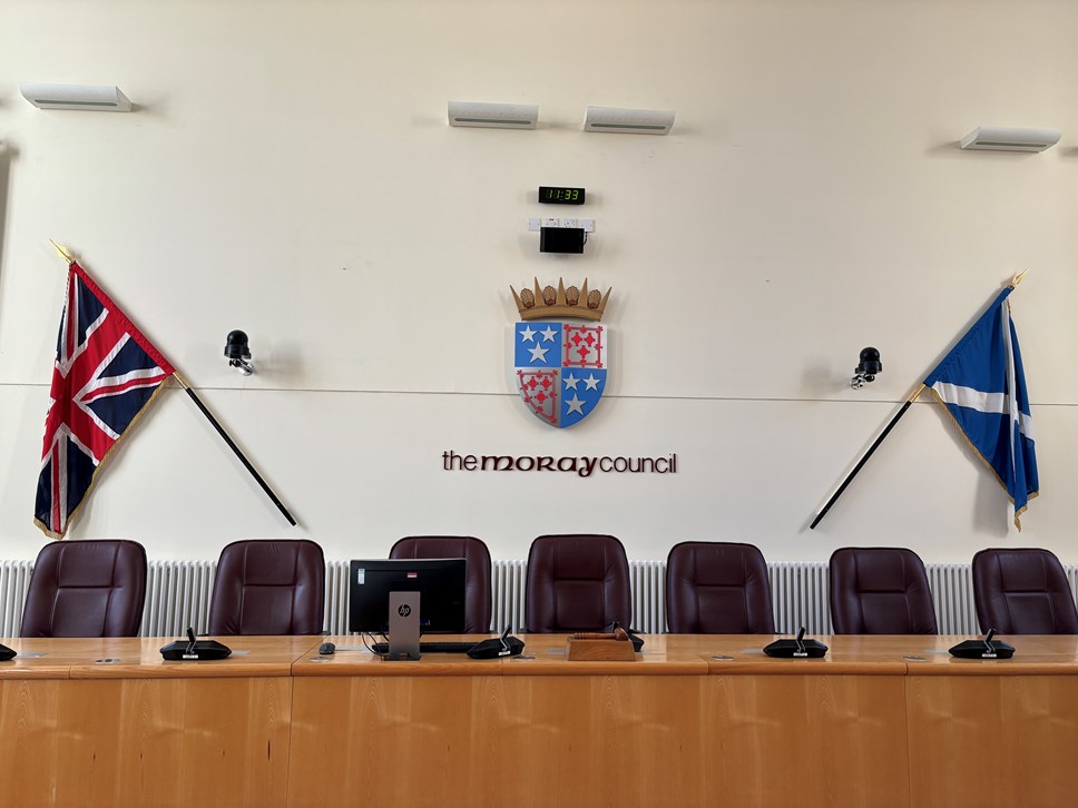 A union flag left, council coat of arms, saltire right with a row of office chairs in front and a wooden desk.