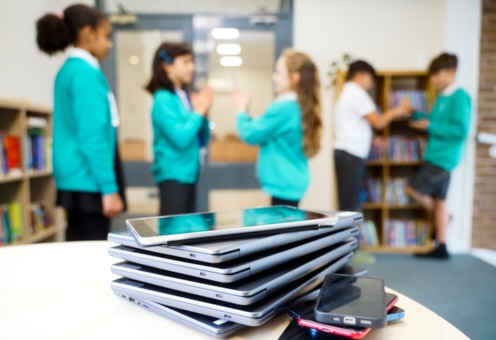 DisconnectToReconnectE: Image shows a close up of various devices, such as ipads, laptops and phones, on a table with children playing in the background.