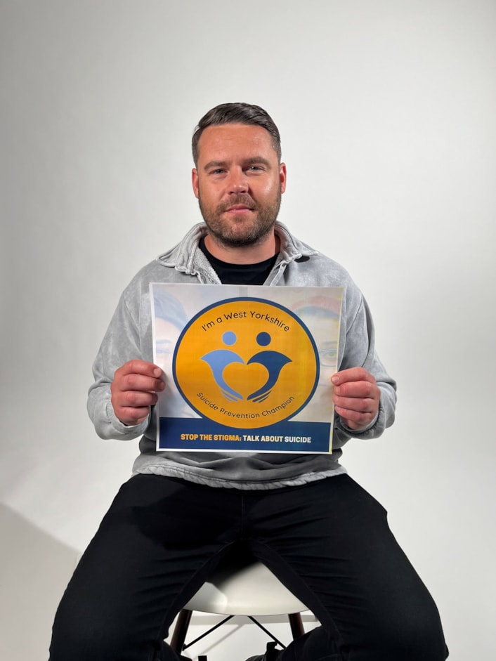 Suicide Prevention Champions - Danny Miller: Danny is pictured sitting on a stool, looking at the camera, holding the Suicide Prevention Champion logo