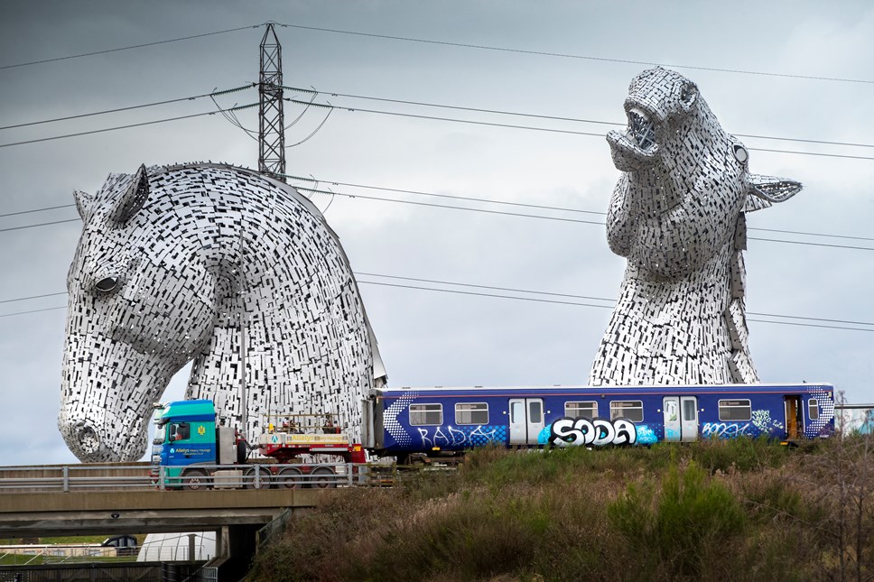 Image of Scotrail carriage loaded onto a truck, on its way to Bo-Ness, travelling past The Kelpies at Grangemouth, Forth & Clyde Canal on 18th Dec 2020. The train has been decommissioned and will be taken to Bo'ness where it will be converted to run on hydrogen.