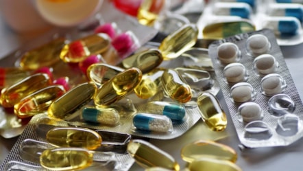 Packets of drugs piled up, some in blister packs, others loose. Pink & white round pills, blue/white and yellow liquid filled oblong pills.