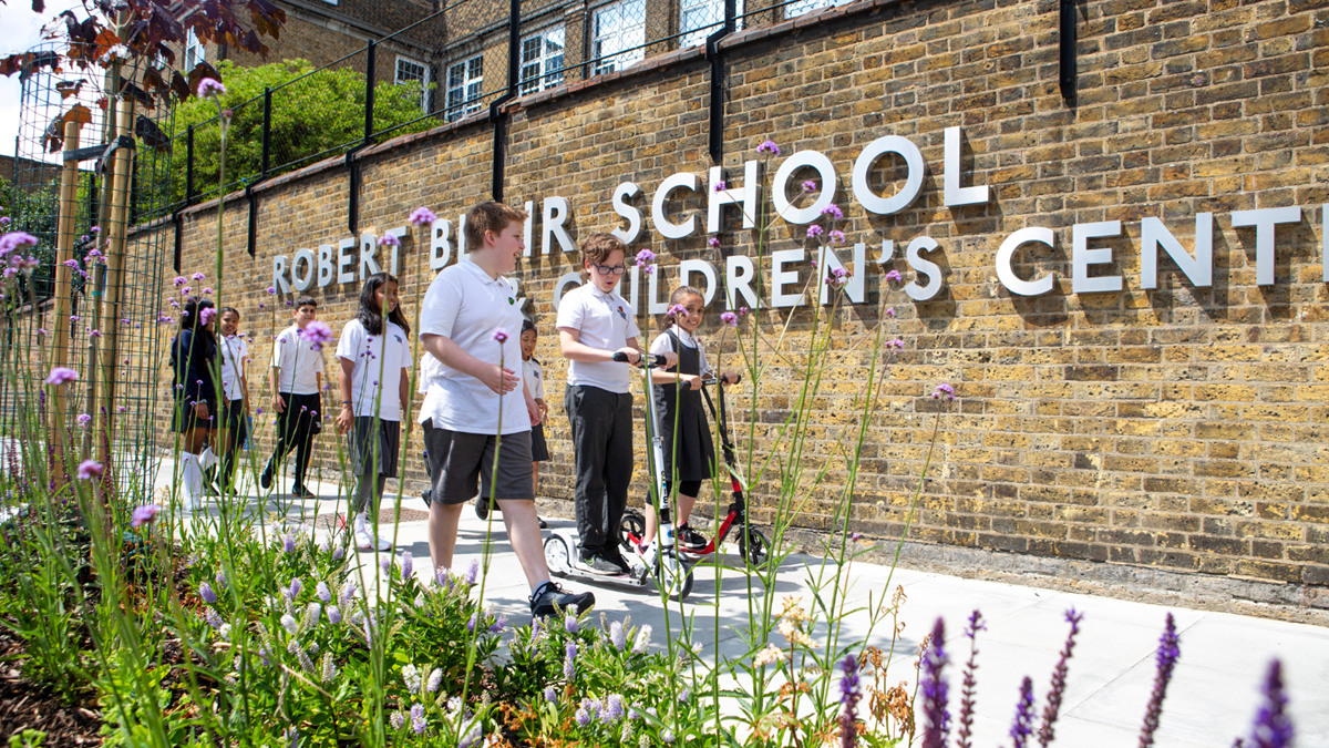 A group of school children in their school uniforms walk and scoot alongside bright green and purple foliage.