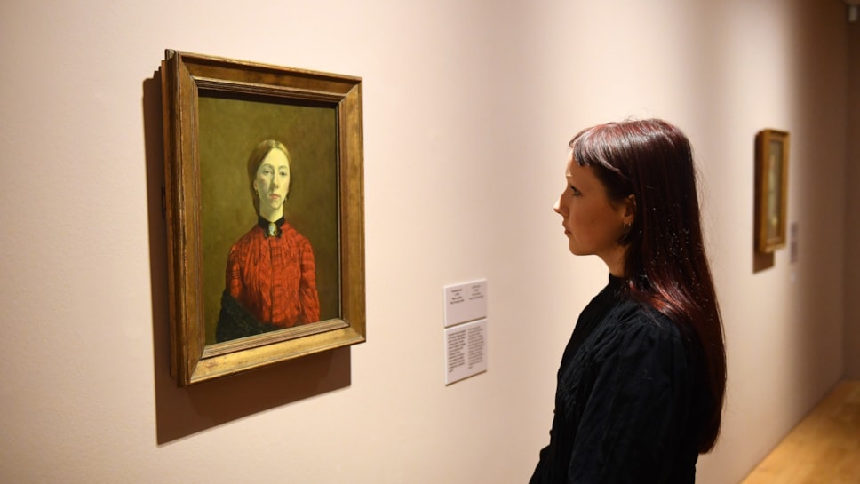 A young woman stands in the Gwen John: Strange Beauties exhibition, studying the painting Self-Portrait, 1902 by Gwen John [Tate]
