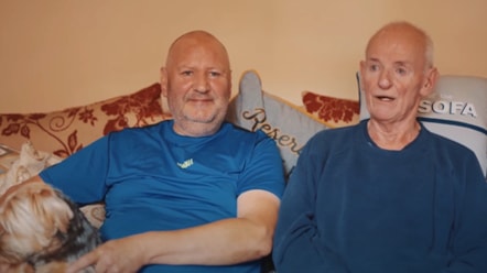 Andrew and John are sitting on a sofa with a small terrier dog. Both men are wearing blue tops. Andrew is smiling.