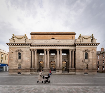 A photograph of the outside of Perth museum, a grand sandstone building with a large open space in front of it. There are people walking in front of the museum in the photo.