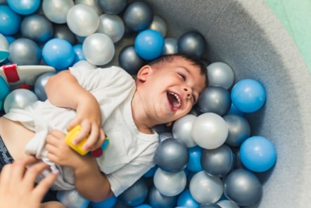 Lewis has a big smile on his face and is wearing a white t shirt and lying on his back in a ball pool with blue, silver and white balls.