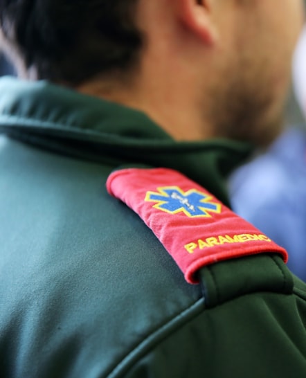 A close up of a green uniform top with a red epaulette bearing the text 'Paramedic', with the blue, yellow and white paramedic logo.