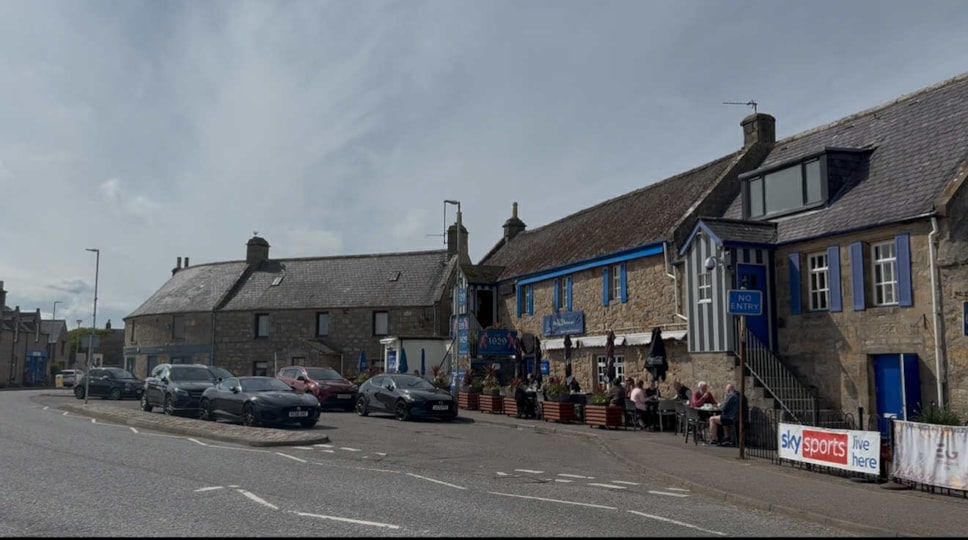 Stone buildings, including a blue trim café and bar; people dine outside near parked cars under a cloudy sky.