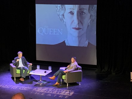 Two men seated on a theatre stage in front of a large projector screen showing a picture of 'The Queen' film of Dame Helen Mirren