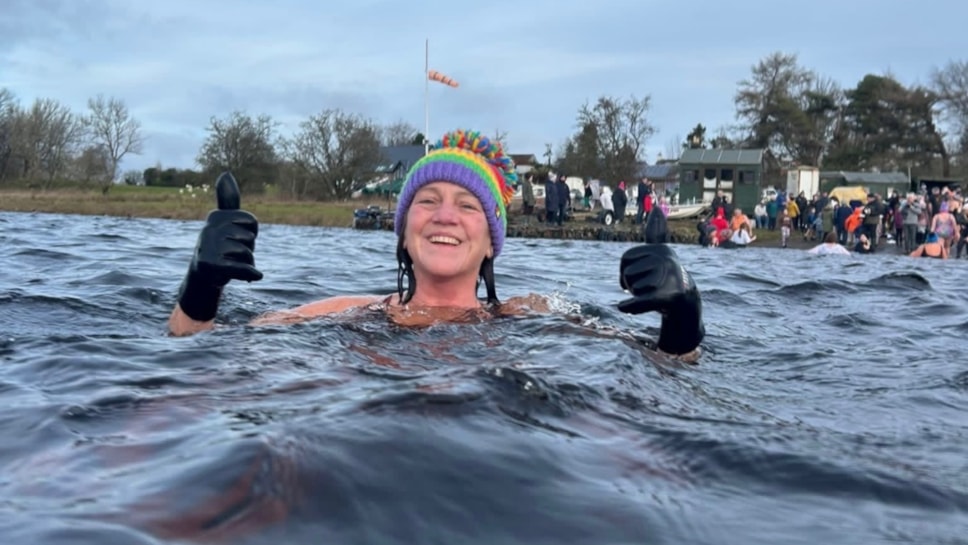Woman in water wearing a multicoloured bobble hat and swimming gloves. She's smiling at the camera