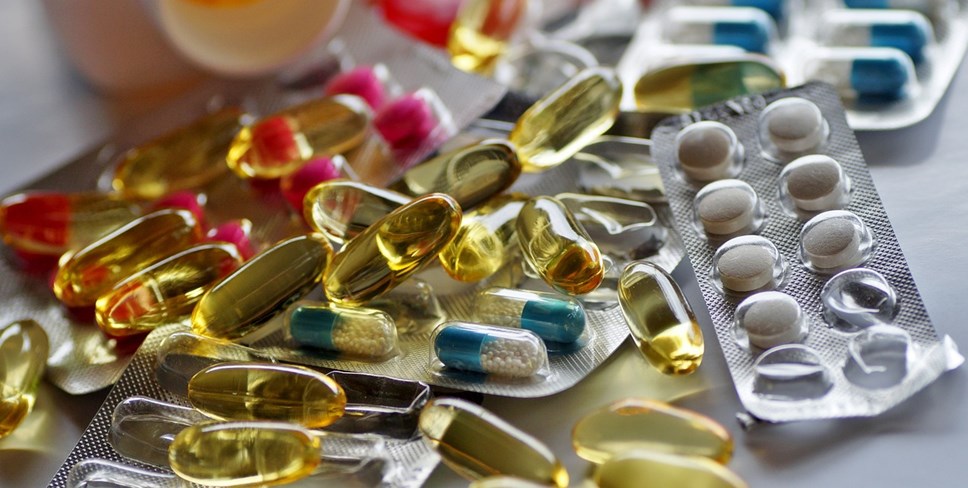 Packets of drugs piled up on a desk. Some are in blister packs, some are loose. Pink and white round pills, blue/white and yellow liquid filled oblong pills.