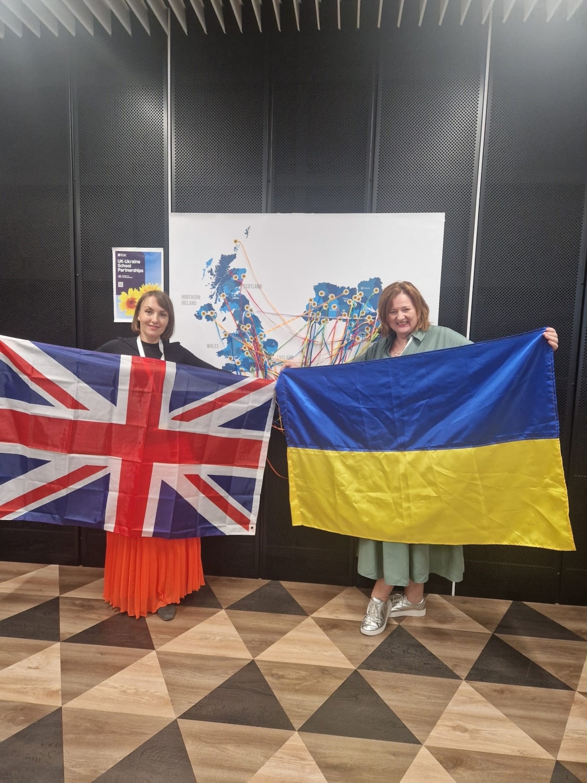 Mrs Toonen, librarian at Elgin Academy, holds the Ukrainian flag, while Ukrainian teacher Katia holds the UK flag. They are standing in front of a dark background with a colourful map display behind them, smiling at the camera.