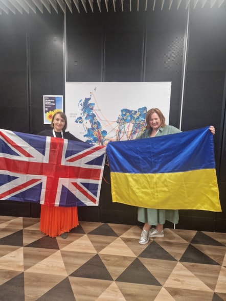 Mrs Toonen, librarian at Elgin Academy, holds the Ukrainian flag, while Ukrainian teacher Katia holds the UK flag. They are standing in front of a dark background with a colourful map display behind them, smiling at the camera.