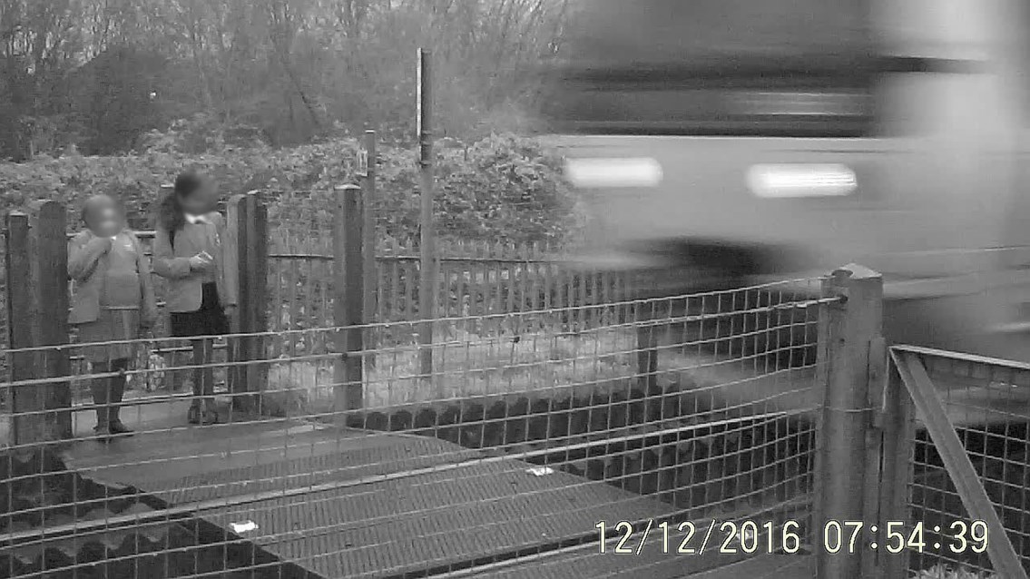 Two school girls stand and wait at Griffin Lane level crossing on the wrong side of the safety gate