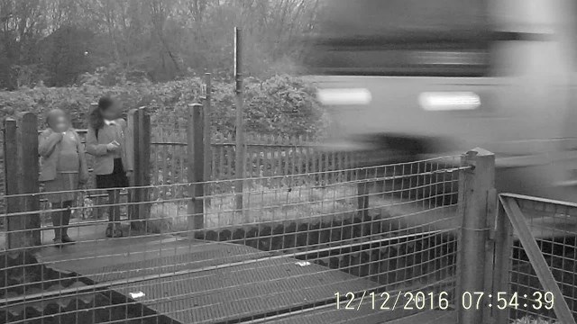 Two school girls stand and wait at Griffin Lane level crossing on the wrong side of the safety gate