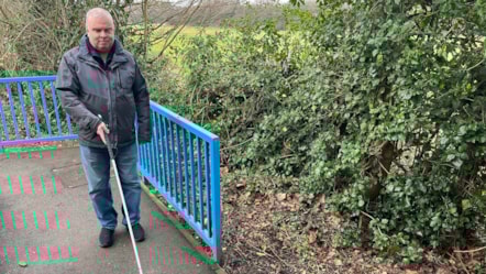 Dave is wearing a grey and black jacket and jeans and is holding a long cane. He's standing in front of blue railings and green leaves