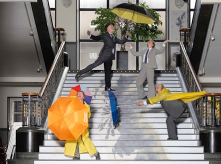 A group of dancers dressed in suits and raincoats post with colourful umbrellas on a set of stairs which has a map of Scotland printed on it